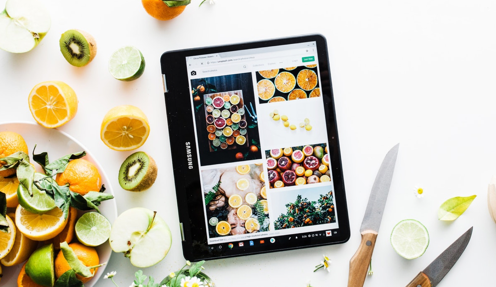Black tablet showing pictures of dishes besides a real dish with vegetables and fruits sitting over a table.