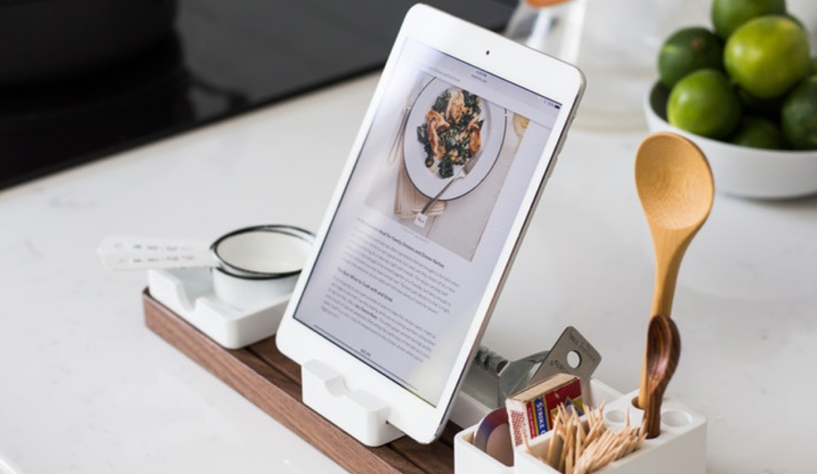 Support holding a tablet displaying a recipe sitting besides wooden spoons and other kitchen utensils.